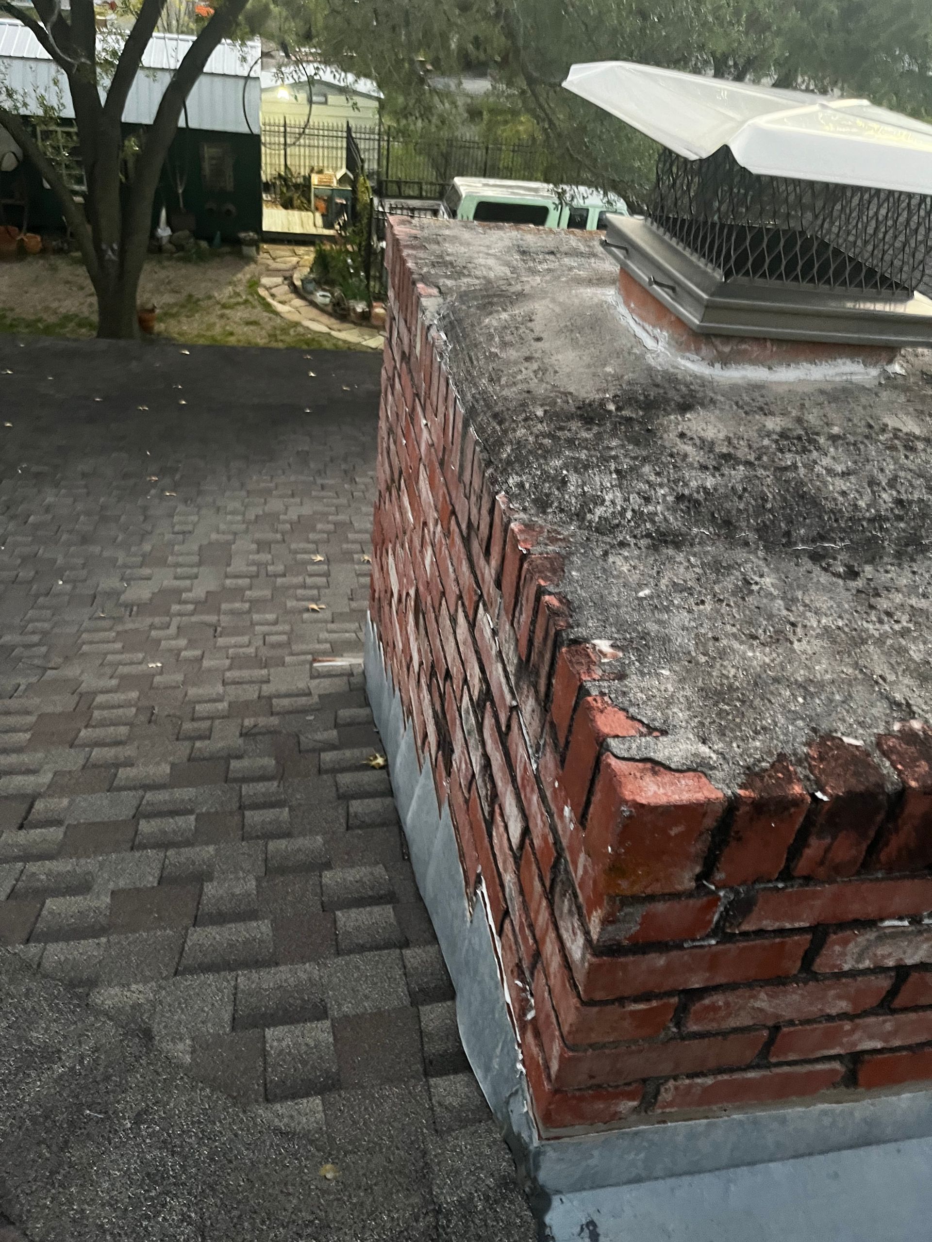 Brick chimney on a shingled roof, with a metal cap. Gray mortar and surrounding roof.