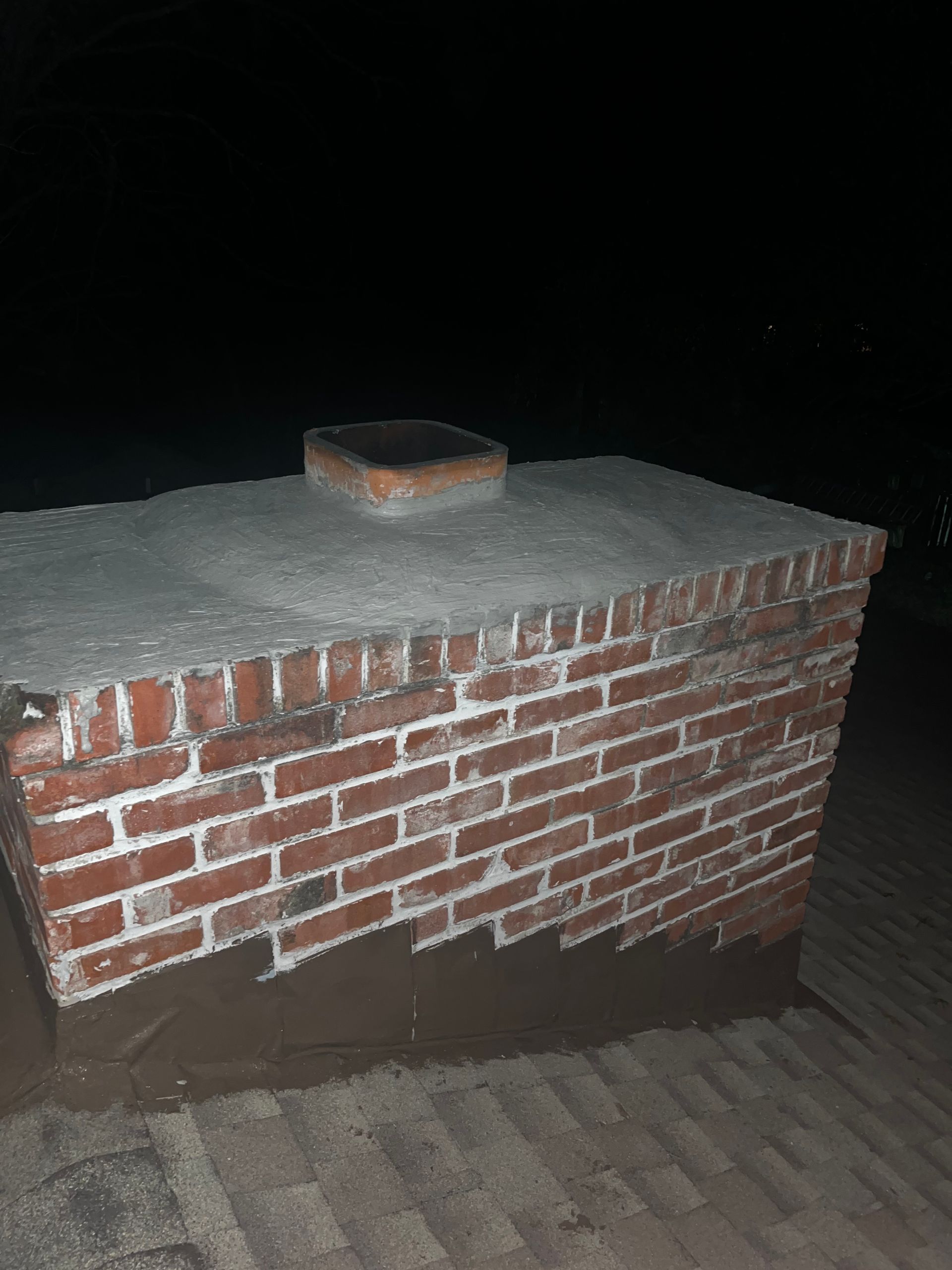 Brick chimney on a roof with a concrete cap and a small flue. The background is dark.