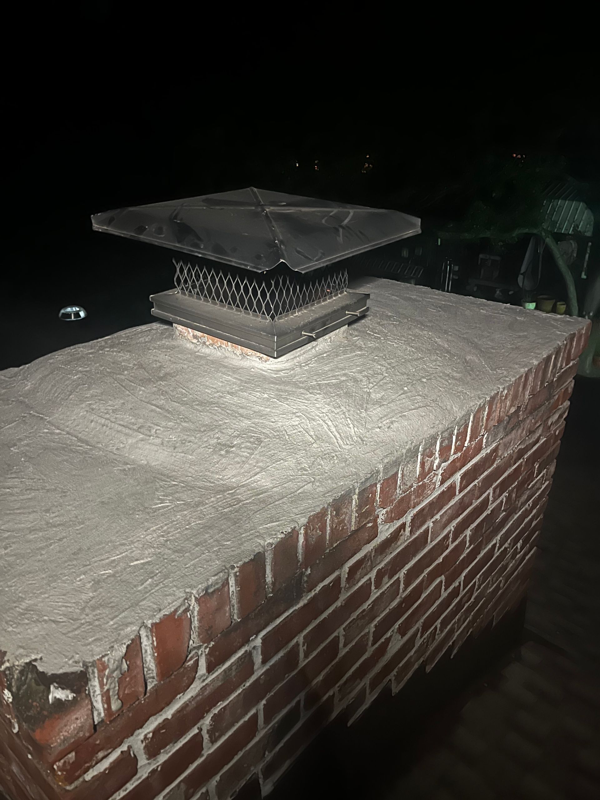 A brick chimney with a black cap, topped with a concrete cap. Dark night setting.