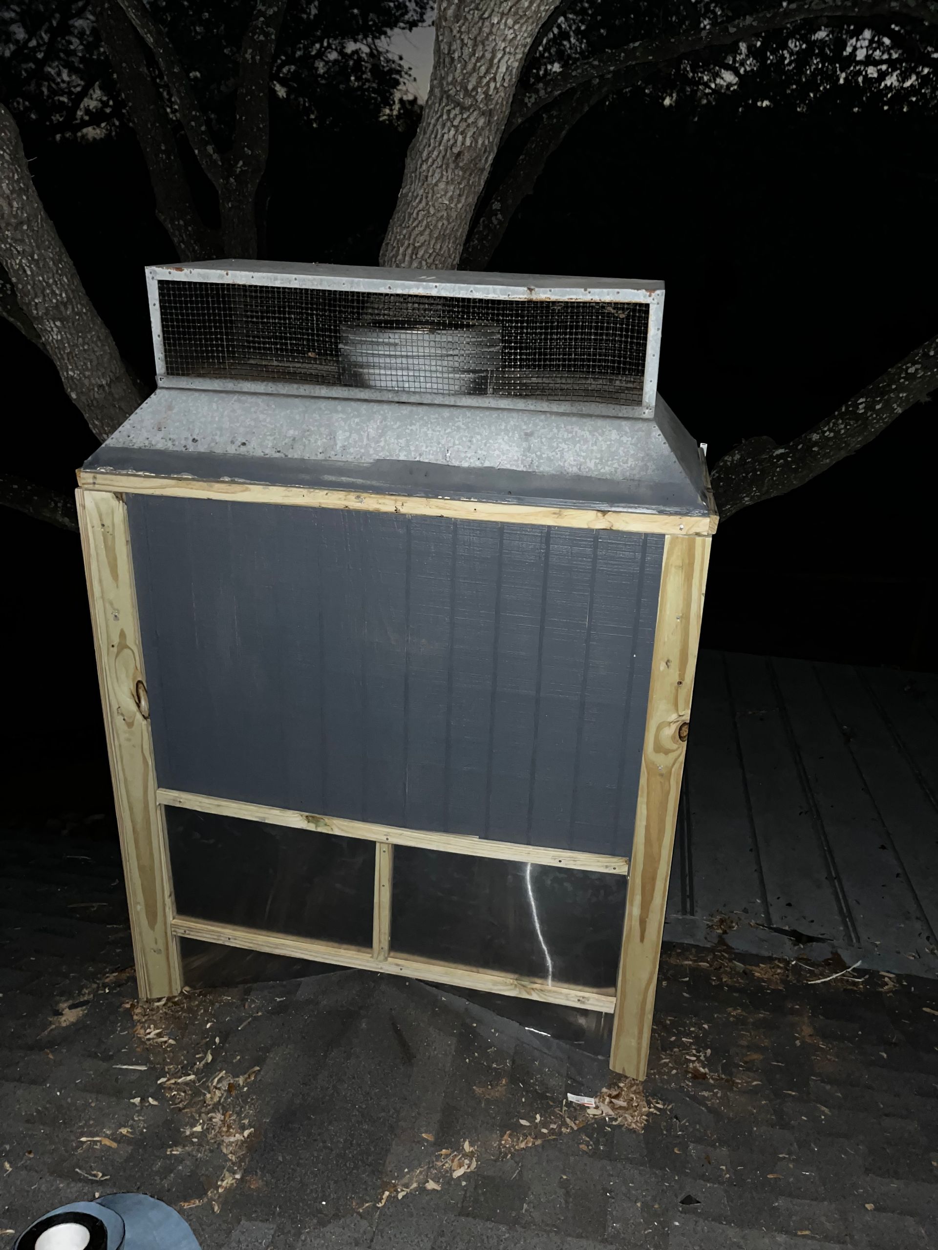Wooden structure with gray front, wire mesh top, near tree at night.