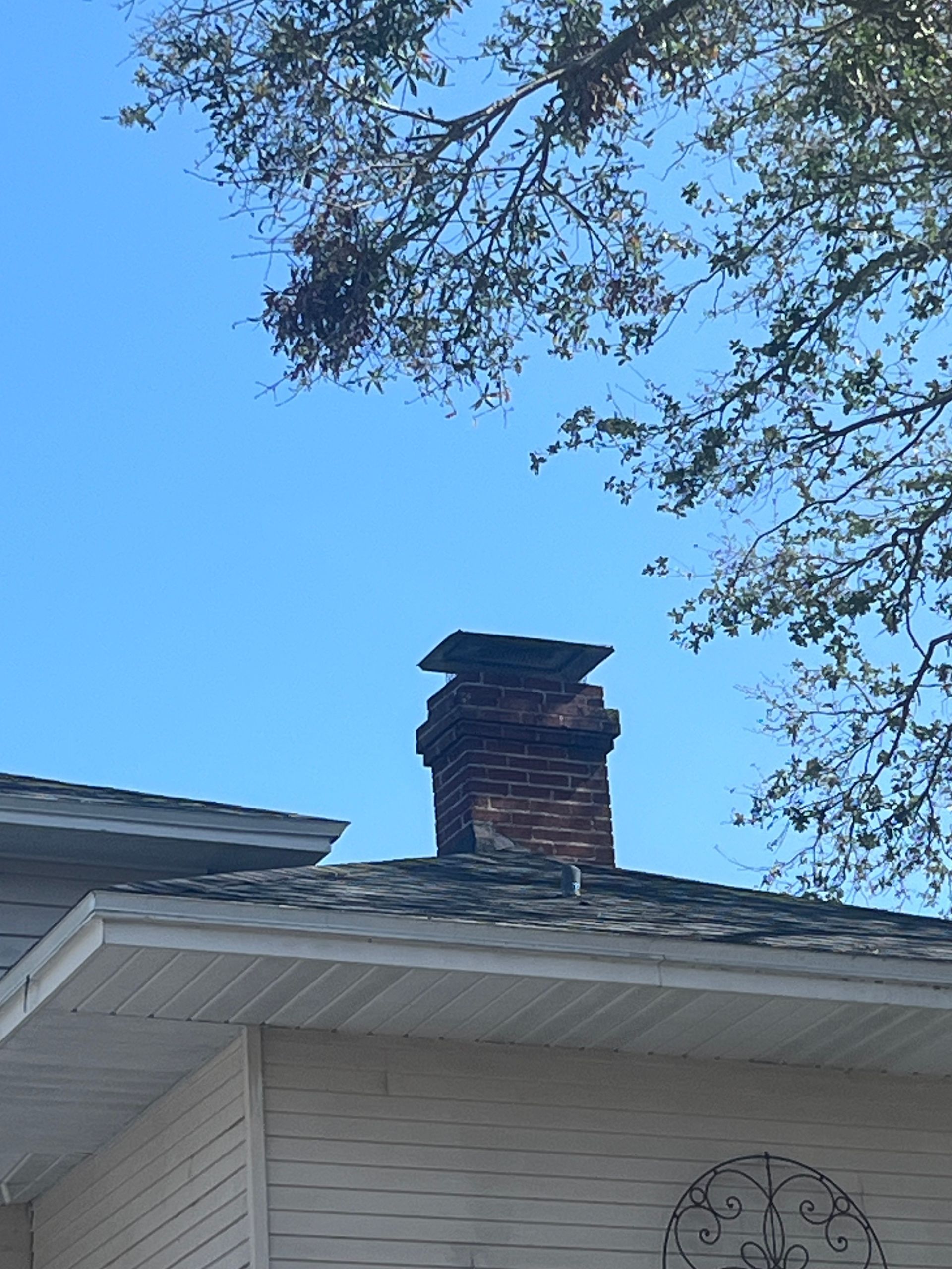 Brick chimney on a shingled roof against a blue sky, with a tree in the background.