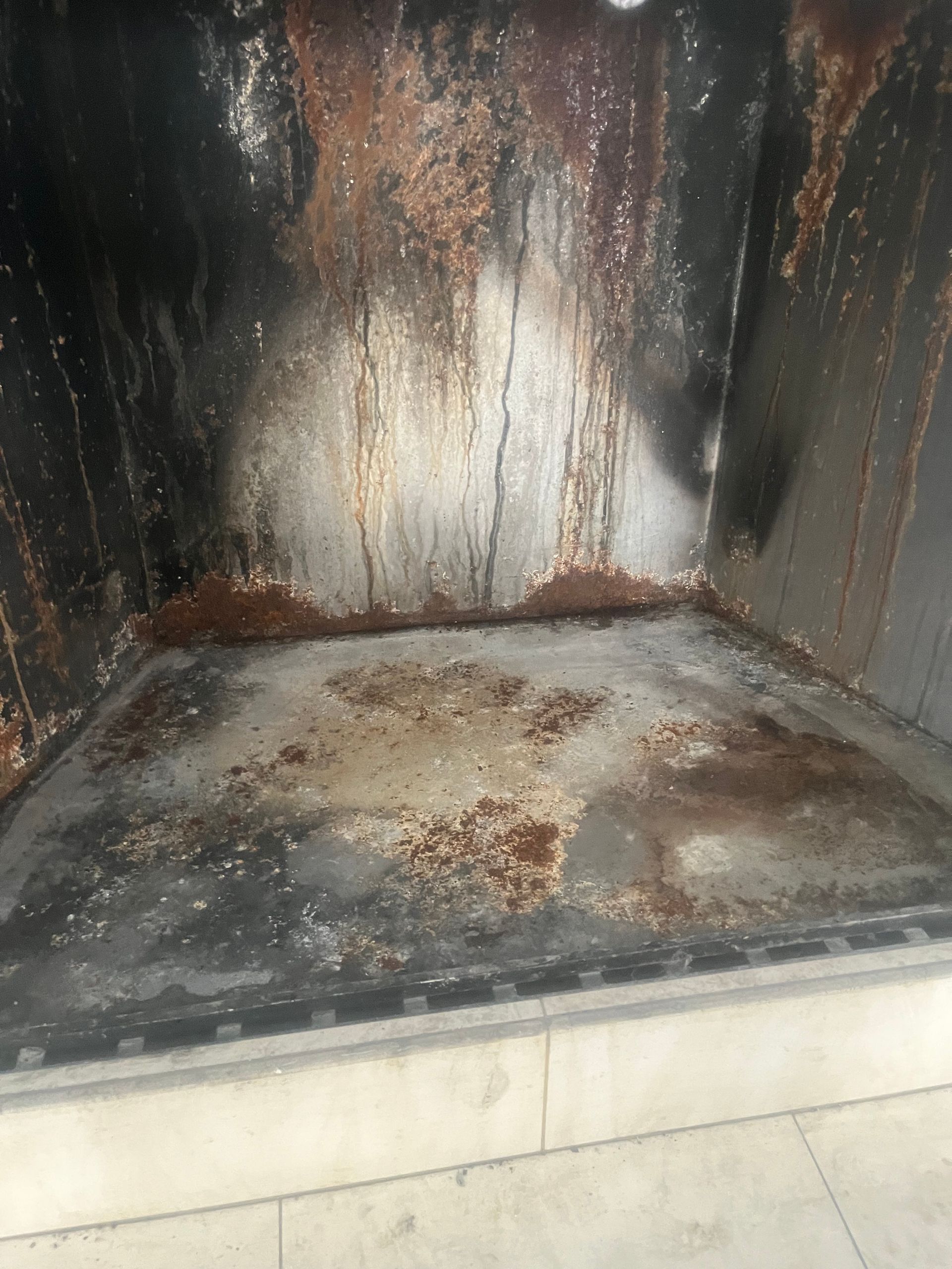 Inside of a dirty, rusty metal fireplace. Brown and black stains cover the walls and floor.