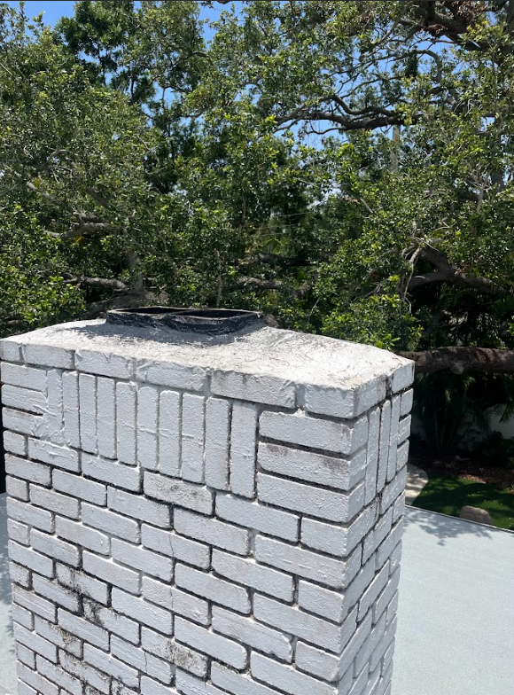 White brick chimney on a rooftop with a metal cap, trees in the background.