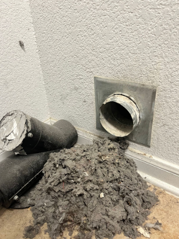 A dryer vent with a large pile of lint, disconnected from the wall duct. Gray walls.