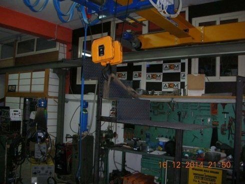 Gru a ponte in un'officina, attrezzature gialle, arancioni e blu, utensili sugli scaffali.