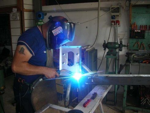 Una persona che salda un metallo in un'officina, indossando un casco da saldatura e guanti, sono visibili scintille blu.