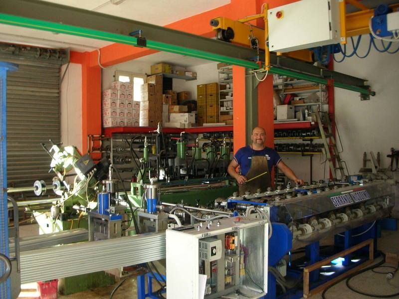 Uomo in officina vicino a una macchina per la formatura dei metalli, gru verde in alto, scatole e utensili visibili.