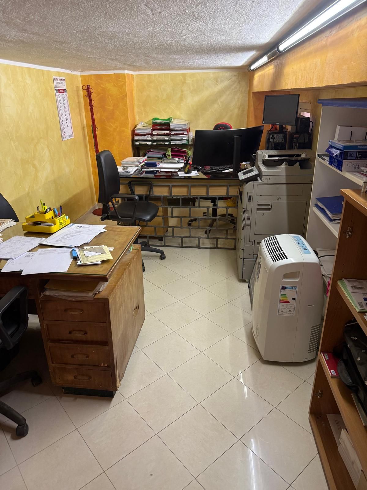 Interno di un ufficio con scrivania, computer, stampante e scaffali. Pareti gialle e pavimento in piastrelle bianche.