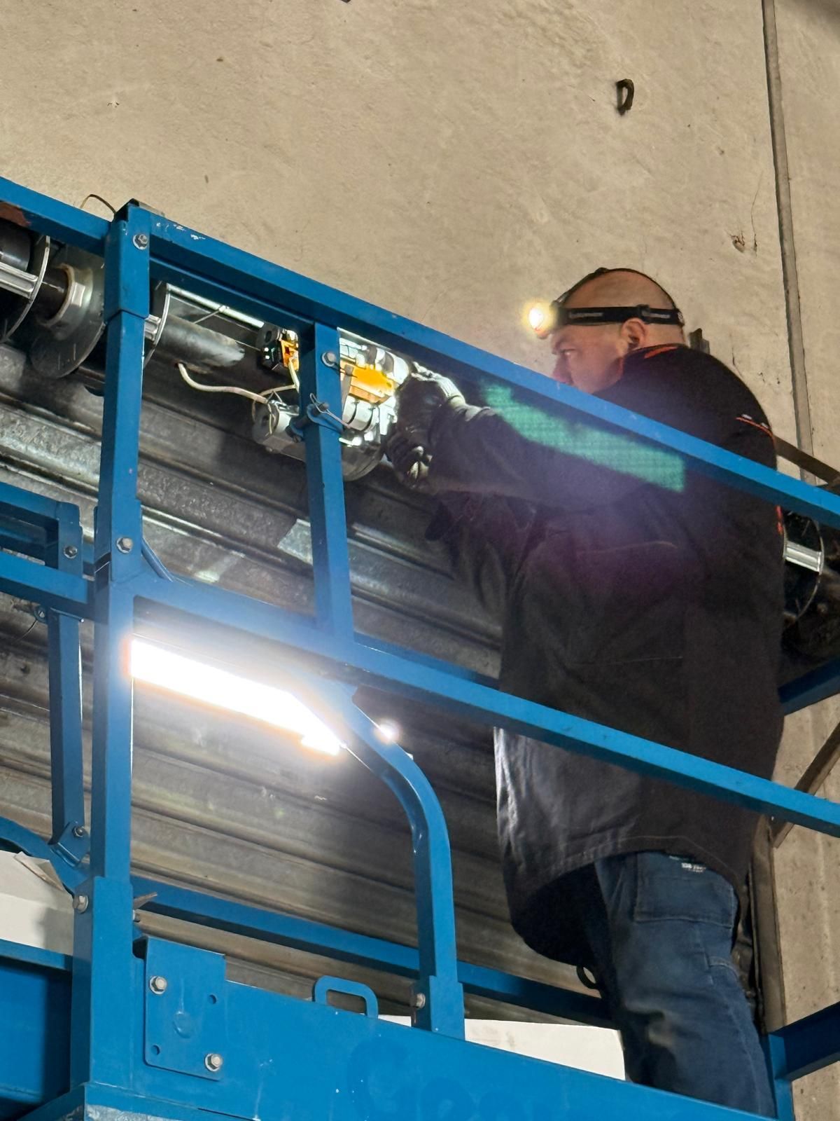 Uomo su ascensore che ripara una saracinesca metallica, con indosso una lampada frontale. Interno industriale, ascensore blu e pareti grigie.