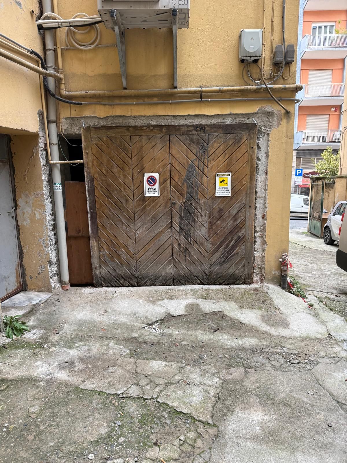 Doppie porte in legno con motivo a zig-zag, due cartelli di avvertimento, inserite in uno stretto ingresso in cemento.