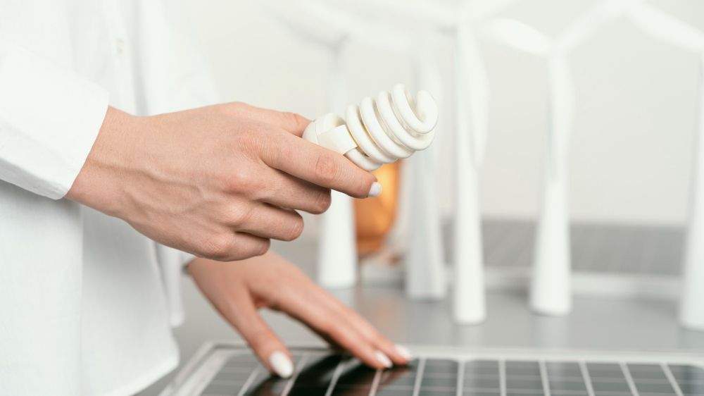 Person holding a lightbulb over a solar panel with miniature wind turbines in the background.
