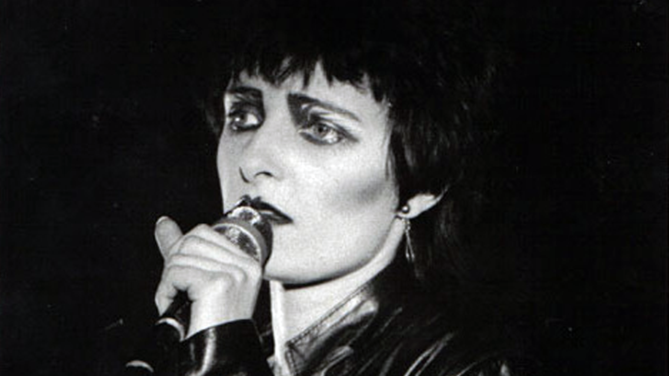 The Post-Punk Origins of Goth Music A black and white photo of Siouxsie Sioux, a key pioneer of goth music, with her iconic dramatic eye makeup and short dark hair, singing into a microphone.