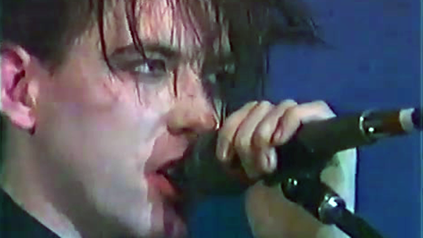 Robert Smith's introspective lyrics and emotive vocals on albums like Pornography defined the sound of early 80s gothic rock. Robert Smith, the frontman of the goth band The Cure, sings passionately into a microphone during a live performance.