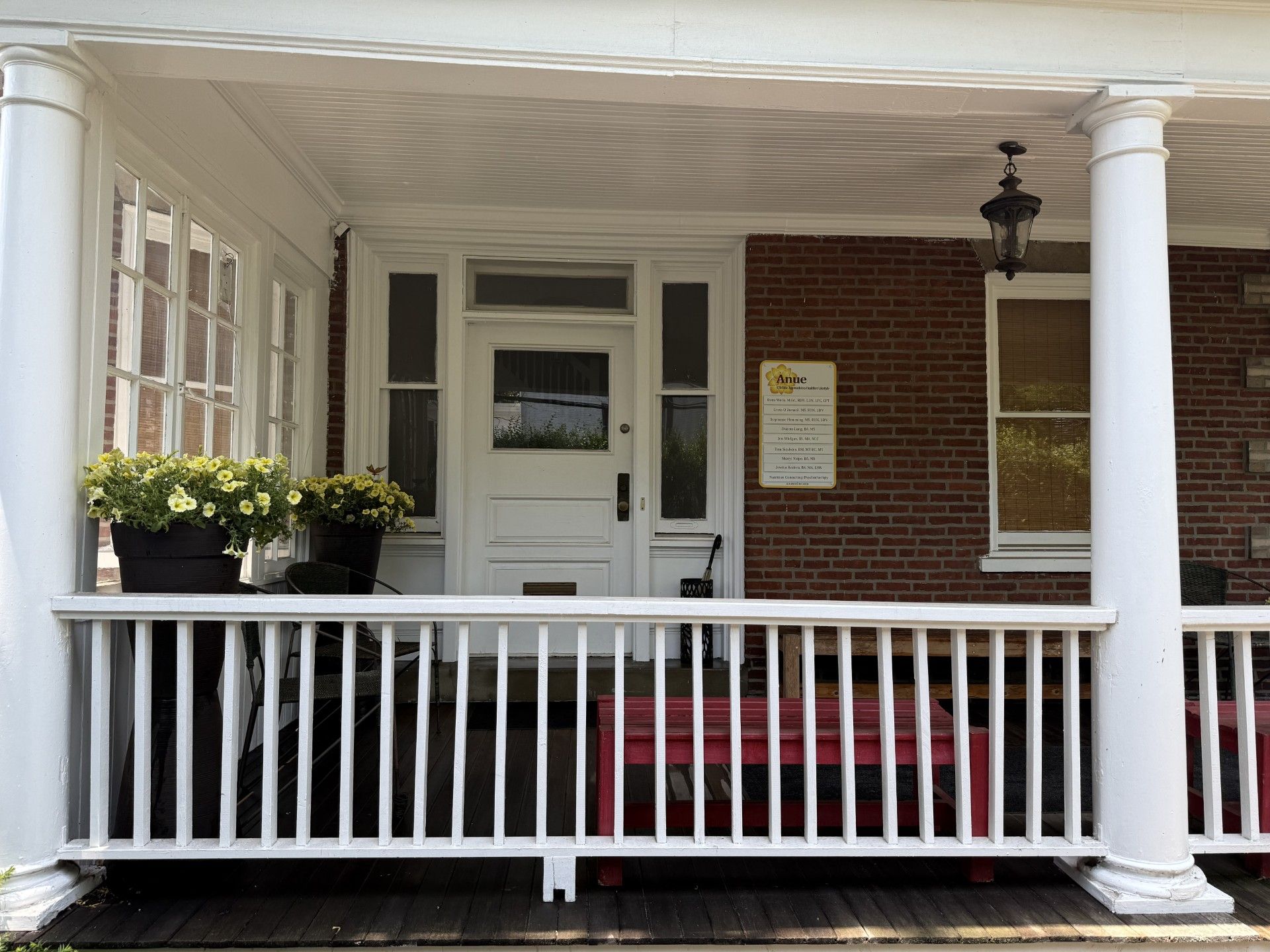White porch of a brick building, with a white door, railing, and potted flowers.