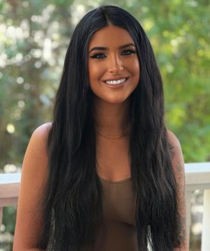 Woman with long black hair, smiling, wearing a tan top, outside with trees in the background.
