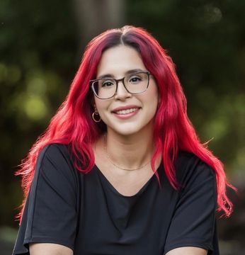 Woman with bright red hair and glasses smiles outdoors.