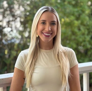 Woman with long blonde hair, smiling, wearing a beige top, on a balcony, green trees in background.