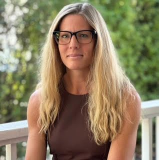 Blonde woman with glasses, brown top, smiles slightly, outside on a porch.