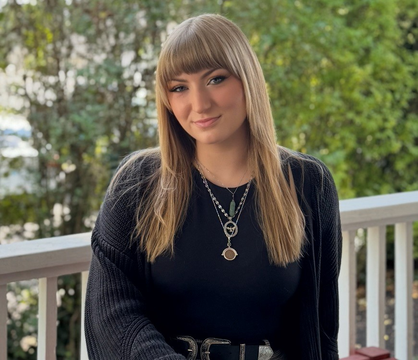 Blonde woman with glasses, brown top, smiles slightly, outside on a porch.