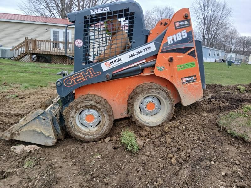 A man is driving a skid steer loader through a dirt field.