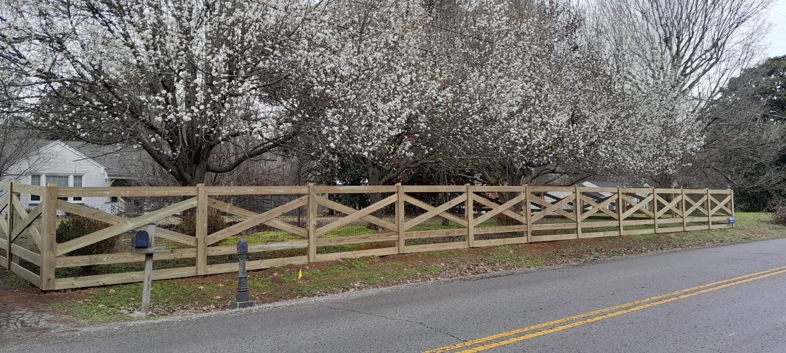A wooden fence with crisscross design in front of a house, with a tree blooming in the background.
