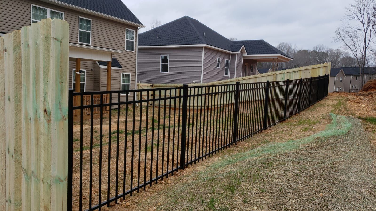 Black metal fence along a dirt path, with new houses in the background. Wooden fence post in foreground.
