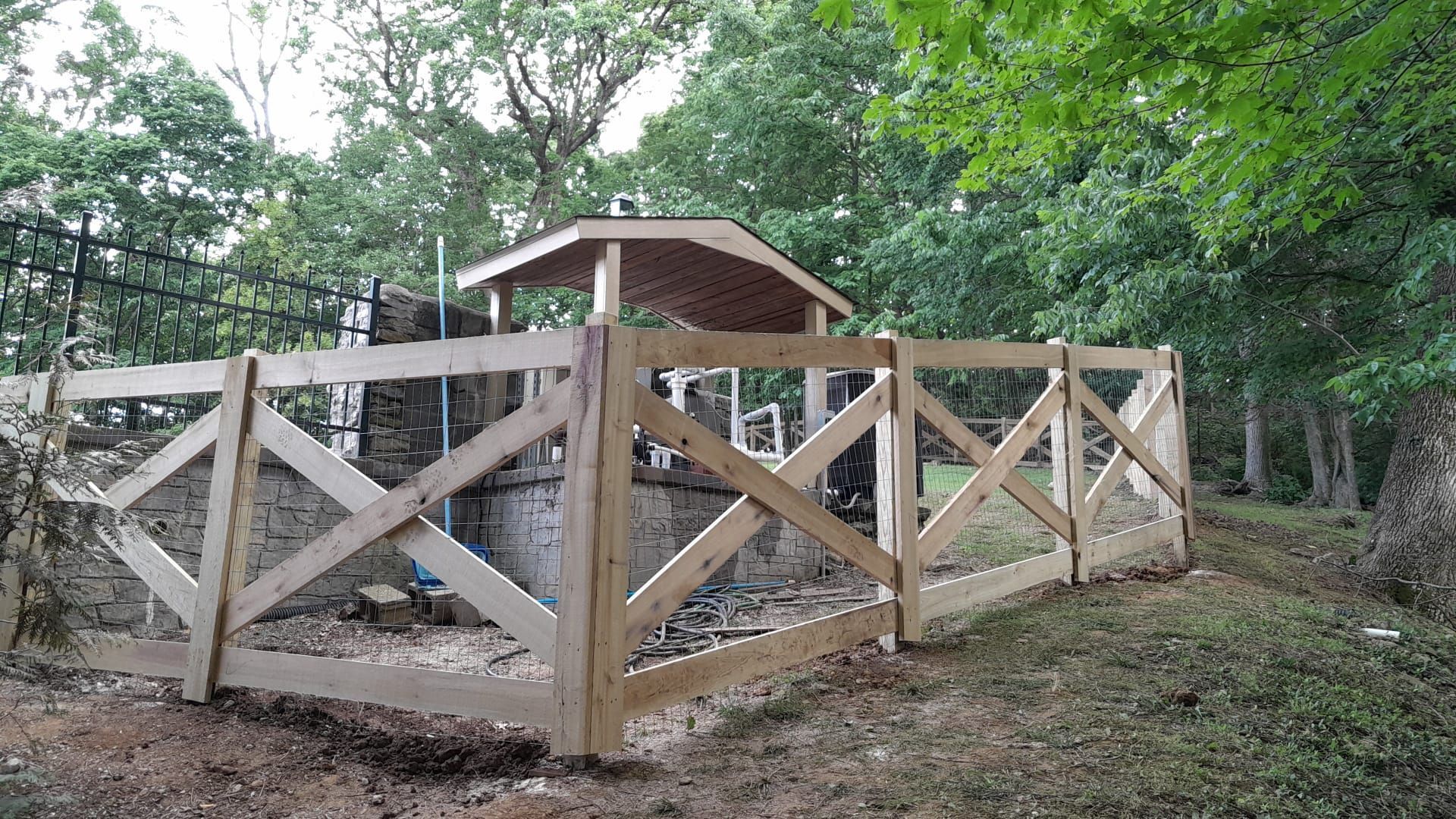 Wooden fence with crisscross design surrounds a small structure with a gazebo-like roof, trees in the background.