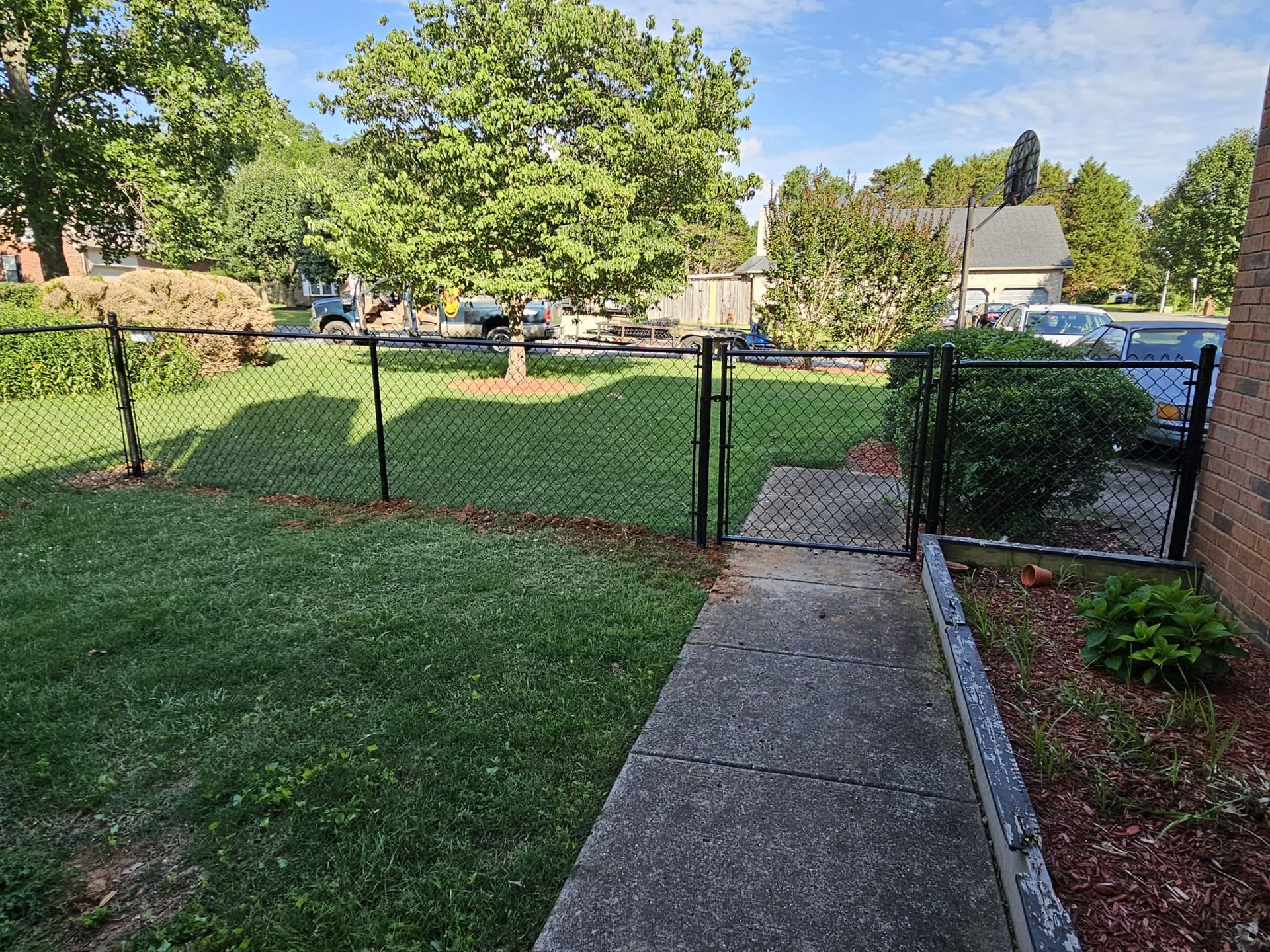 View of a yard with a chain-link fence, concrete pathway, and gate. Green grass and trees are visible.