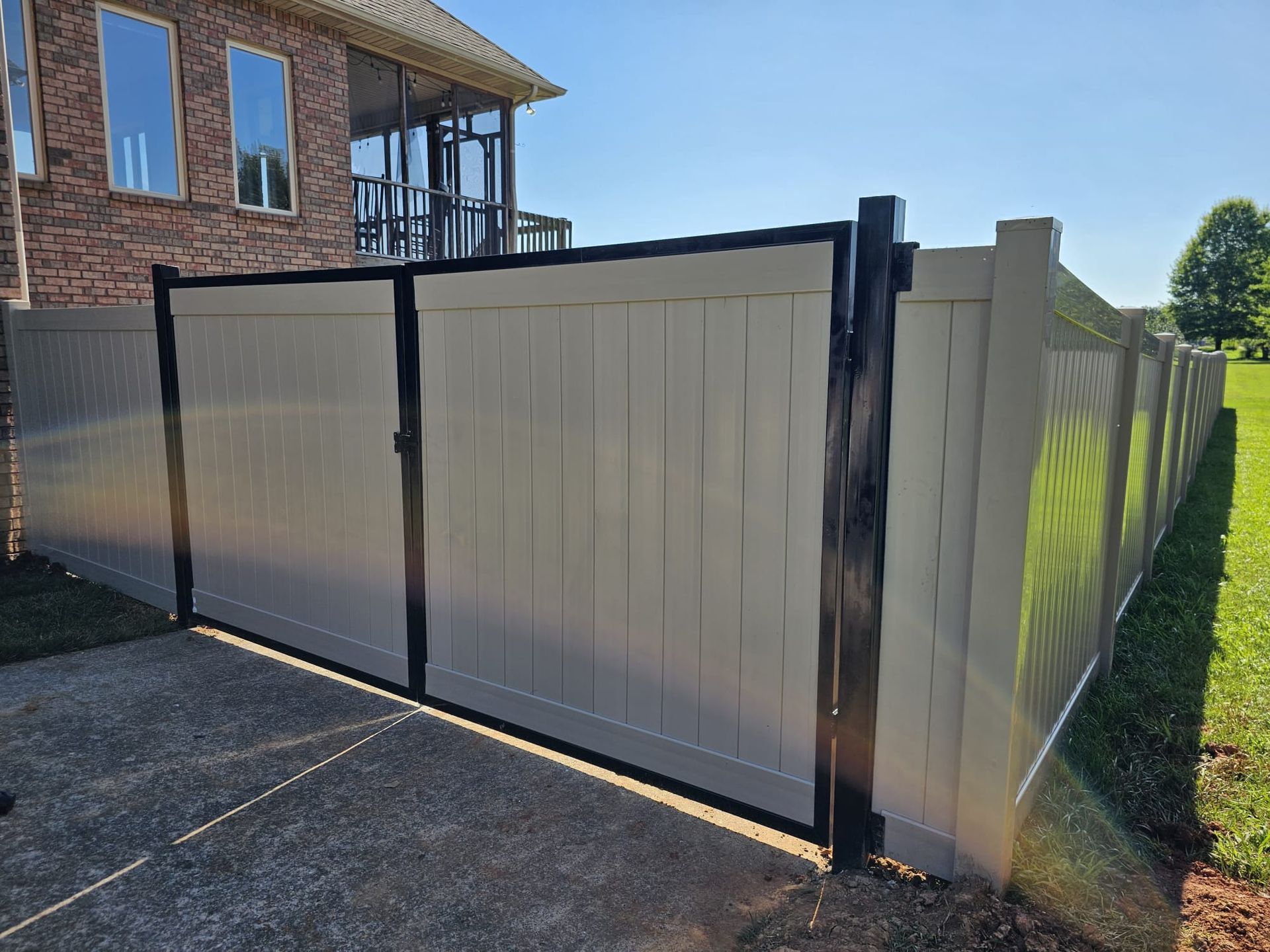 Tan and black vinyl fence with gate, enclosing a backyard on a sunny day.