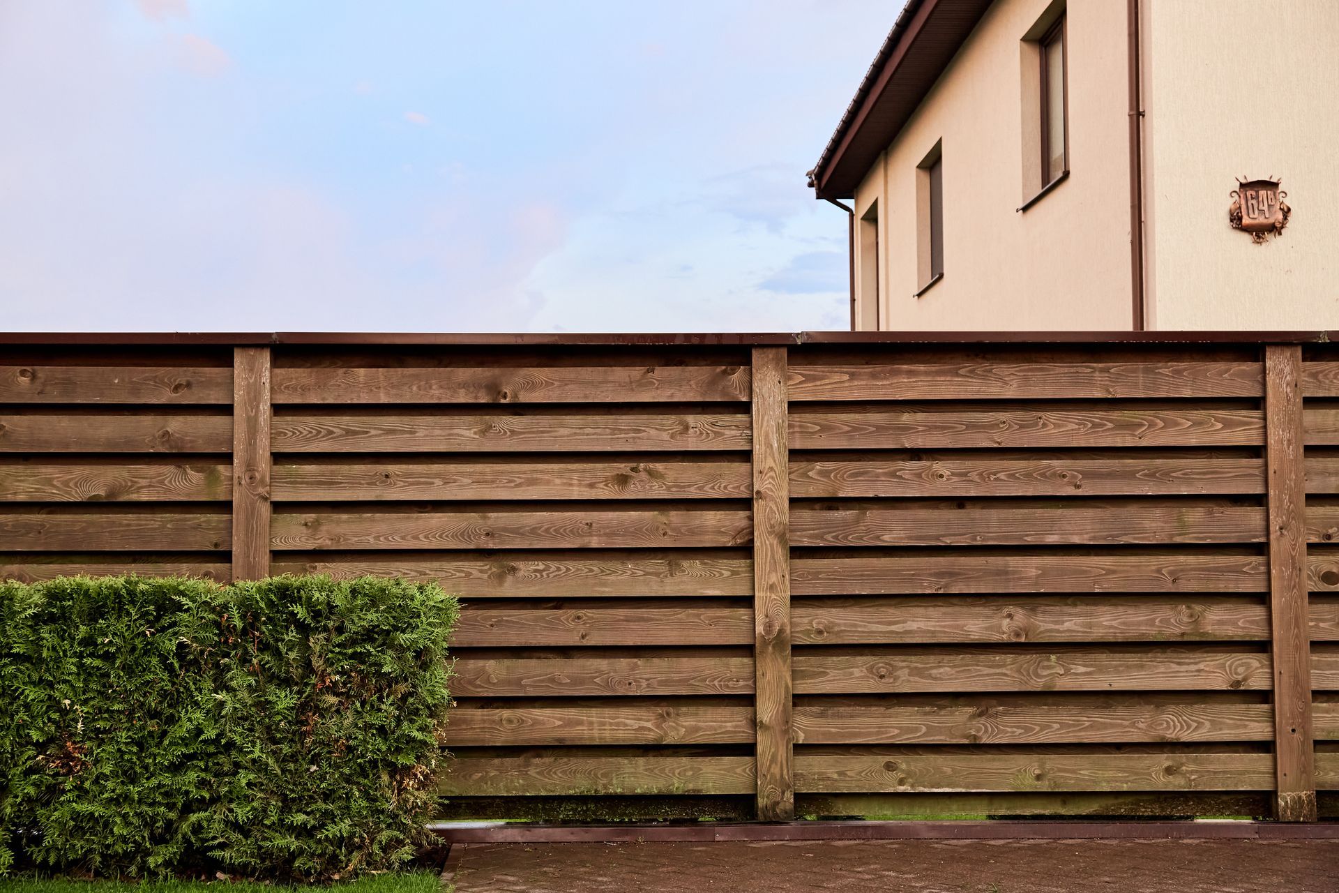Wooden fence with green hedge, beige house in background under cloudy sky.