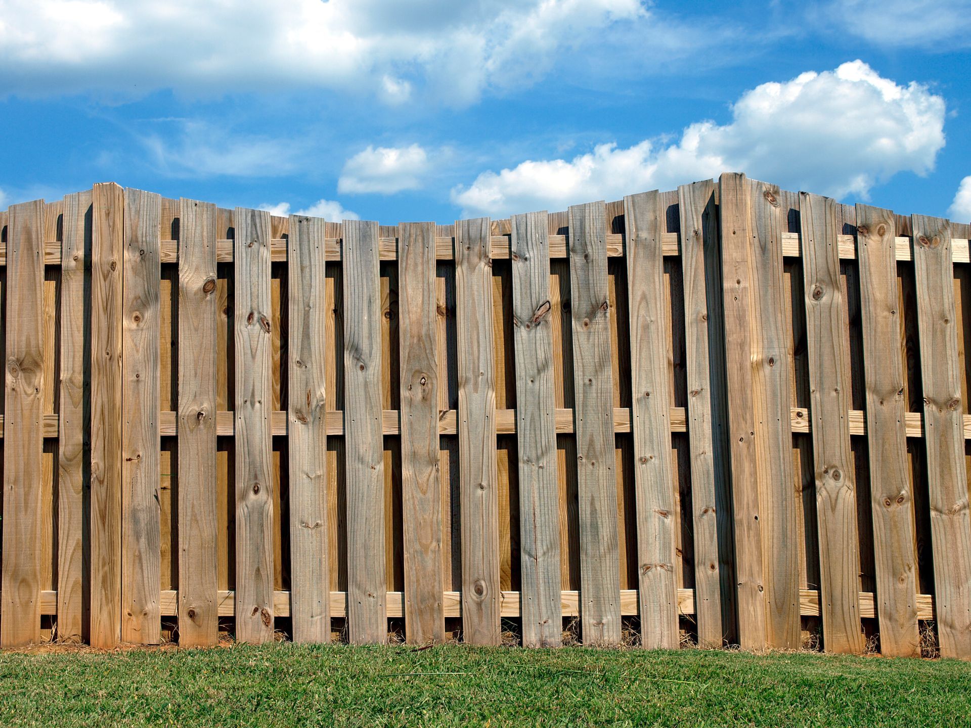 Wooden fence on green grass against a blue sky with fluffy white clouds.