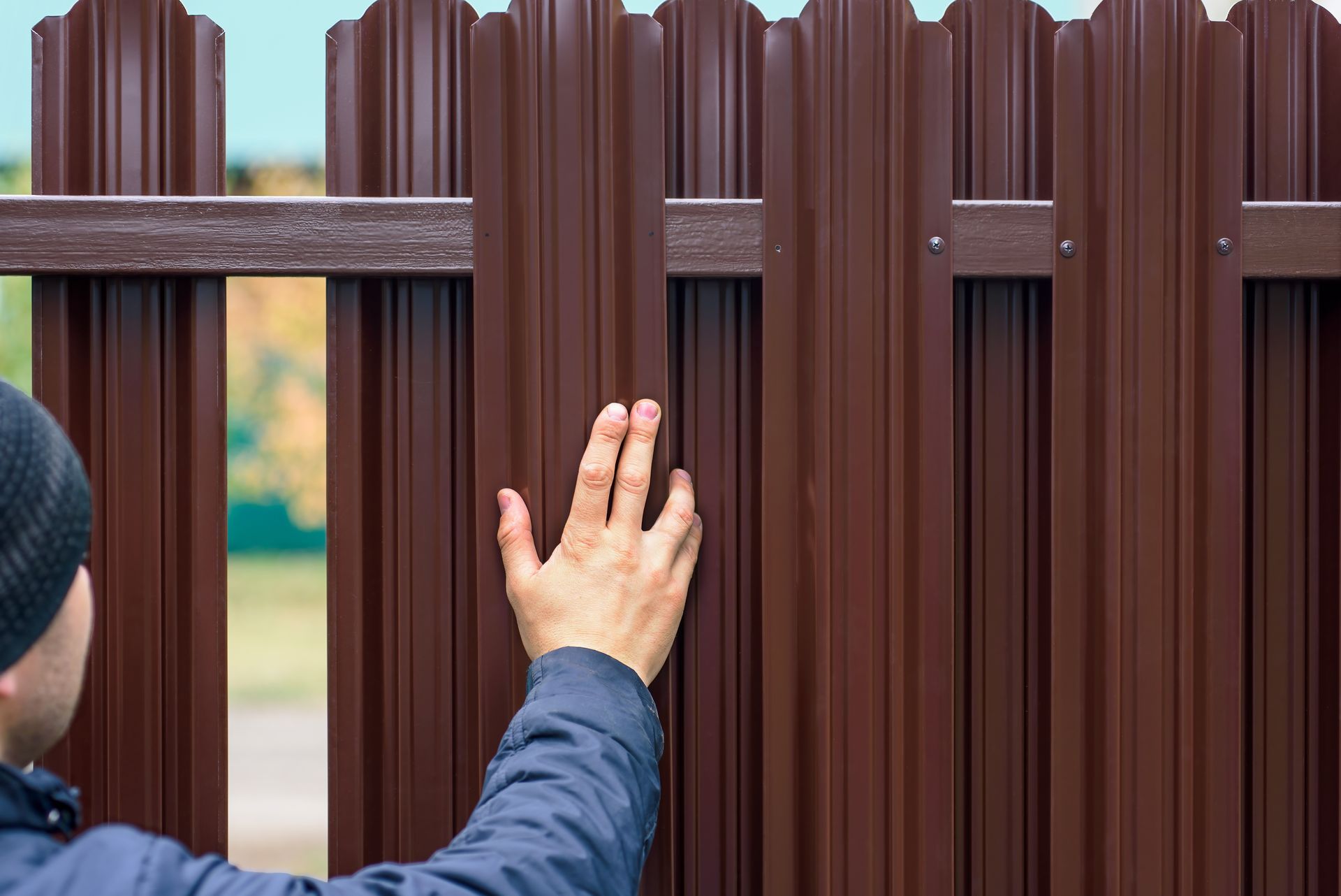 A person in a blue jacket reaching out to touch a brown metal picket fence outdoors.