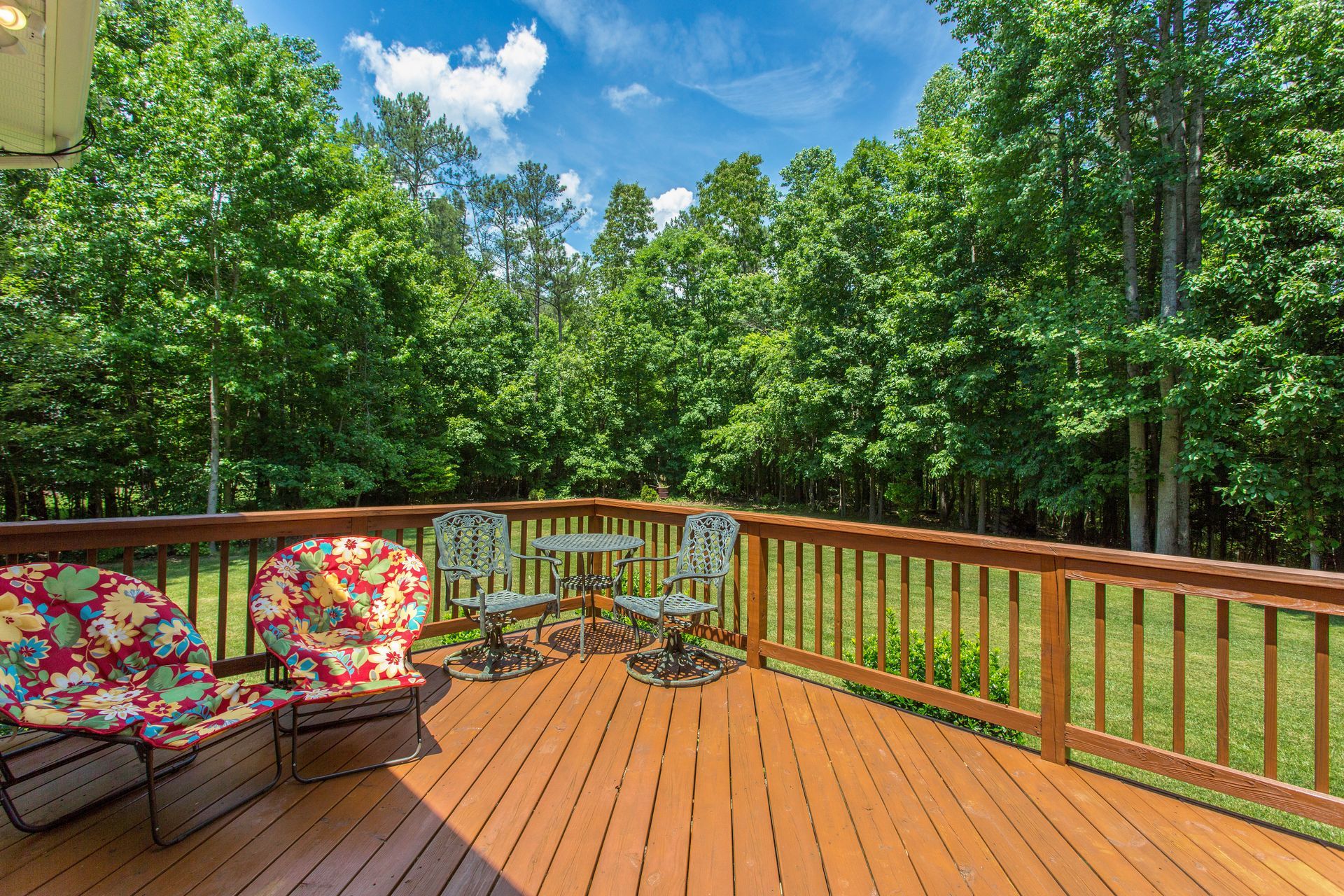 Wooden deck with chairs and table, overlooking a lush green wooded area under a blue sky.