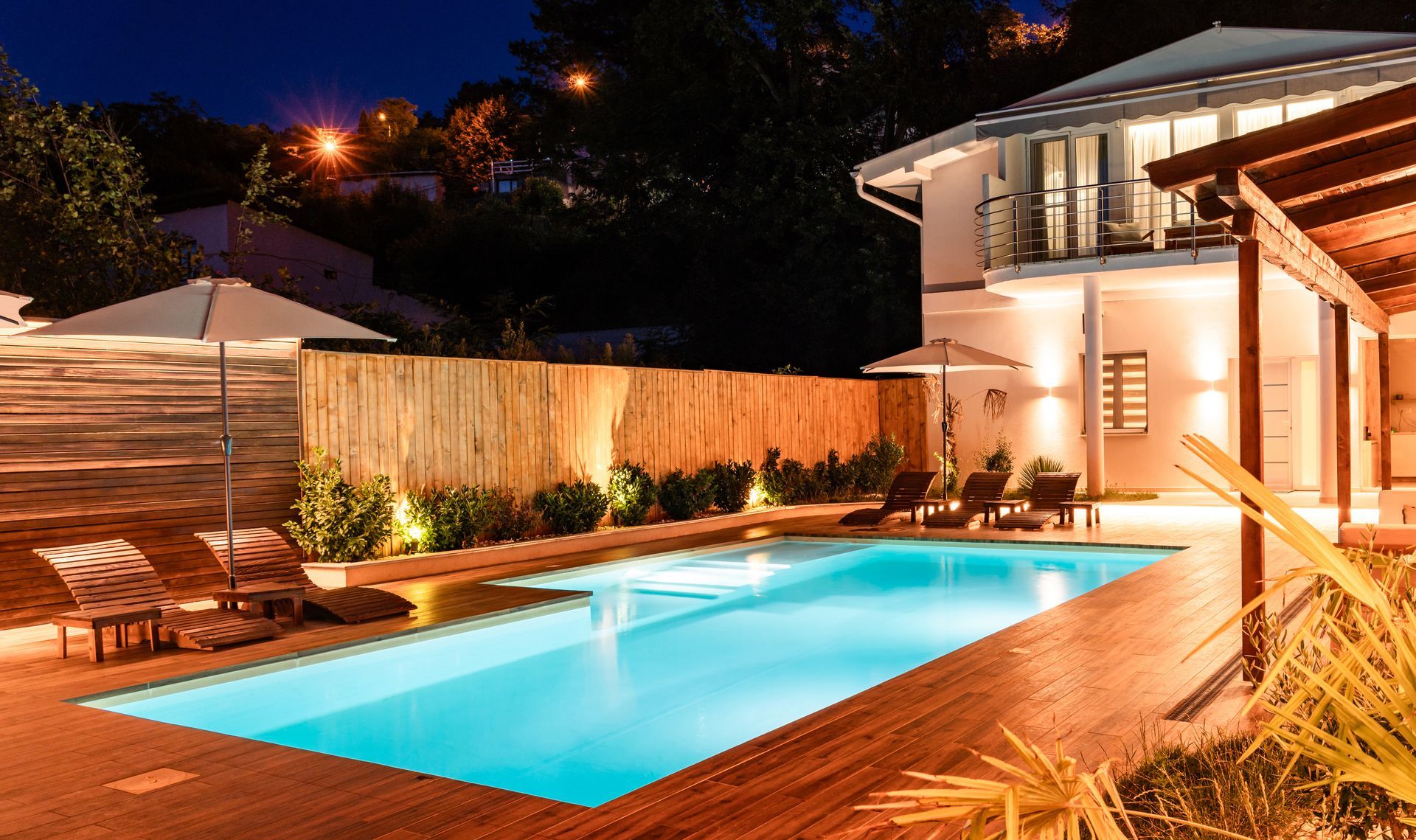 Night view of a pool with a lit house, wooden deck, and lounge chairs.