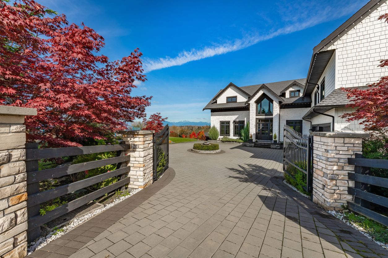 A brick and black gated driveway leads to a white house with a black roof and a blue sky.