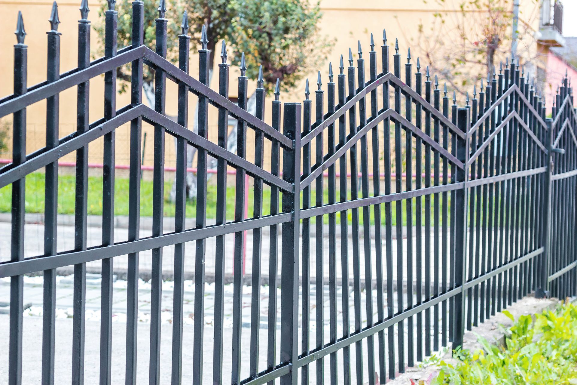 Black metal fence with pointed tops, zigzag top rail, along a sidewalk with green grass.