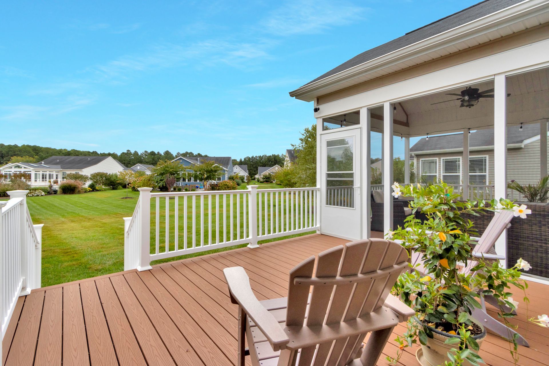 A wooden deck with Adirondack chairs, overlooking a grassy yard with houses under a bright blue sky.