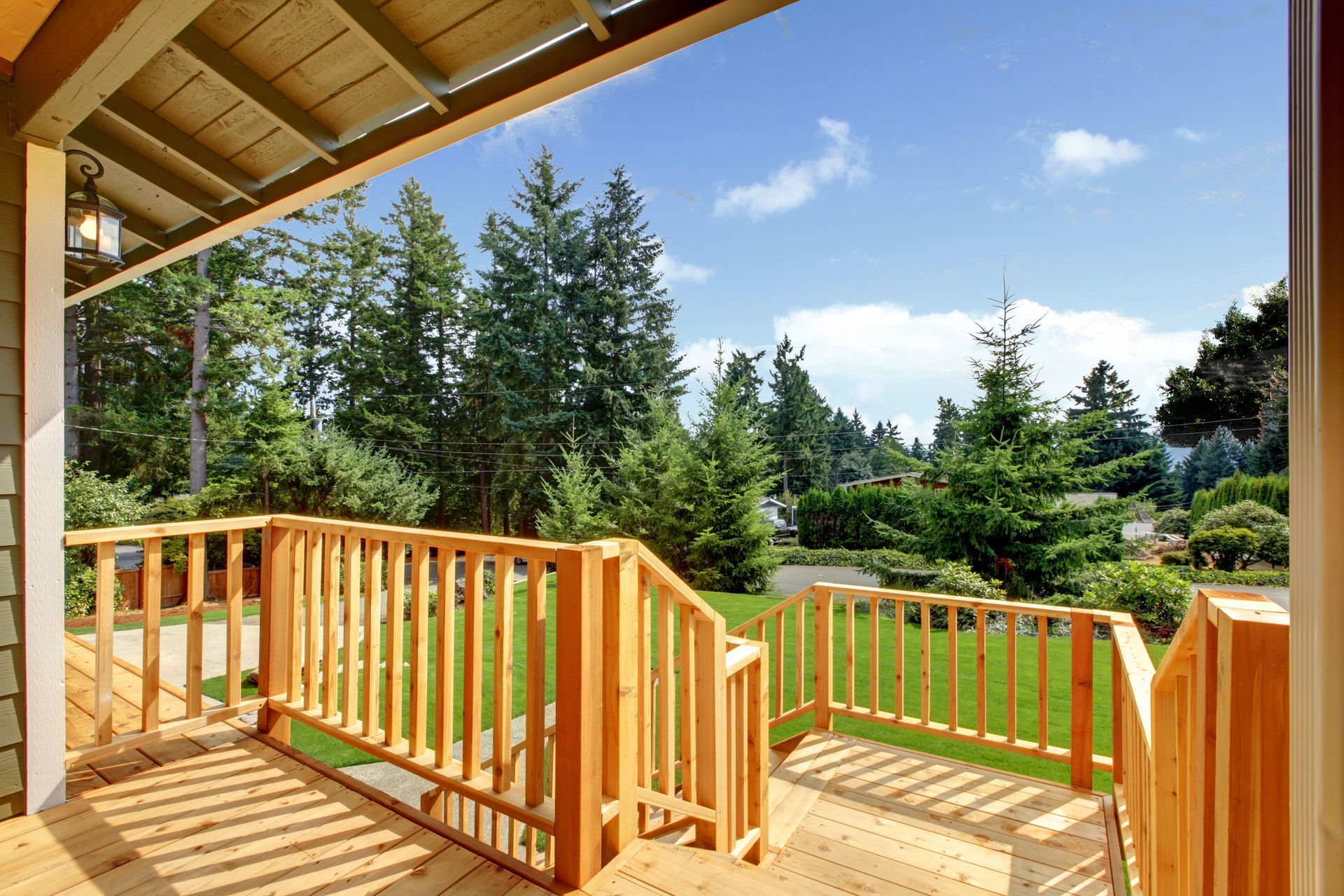 Wooden porch with railing overlooking a grassy yard and evergreen trees under a blue sky.
