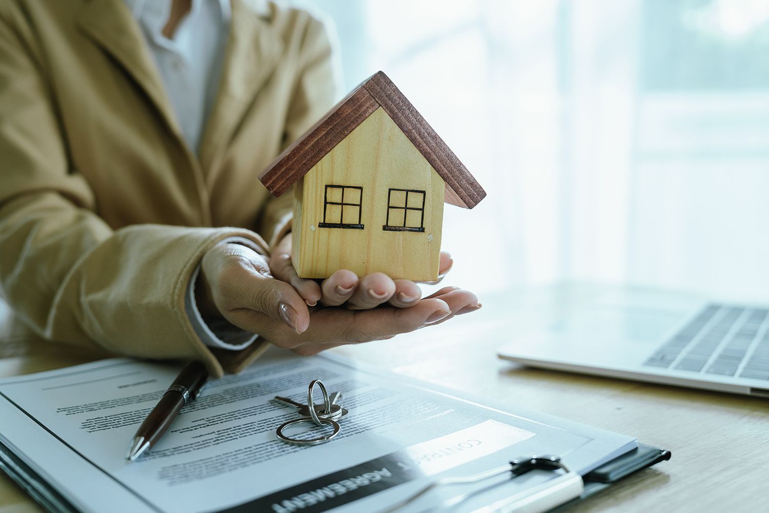 Person in a blazer holds a small wooden house over a contract and keys.