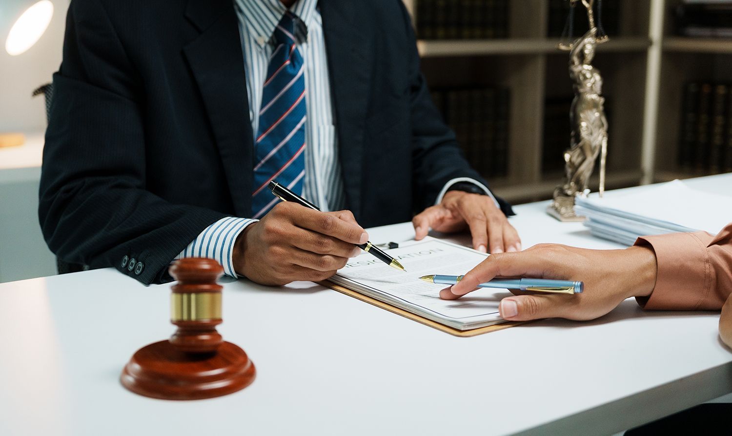 Lawyer in suit signing document with client at desk, gavel in foreground.