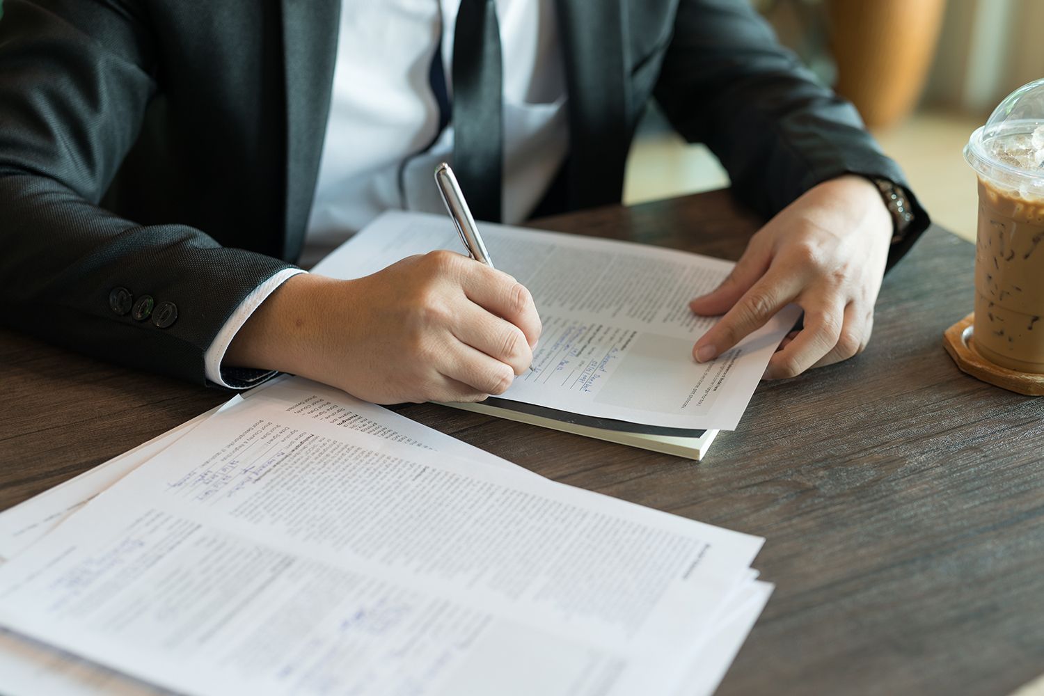 Person in a suit writing on papers at a table with a drink nearby.