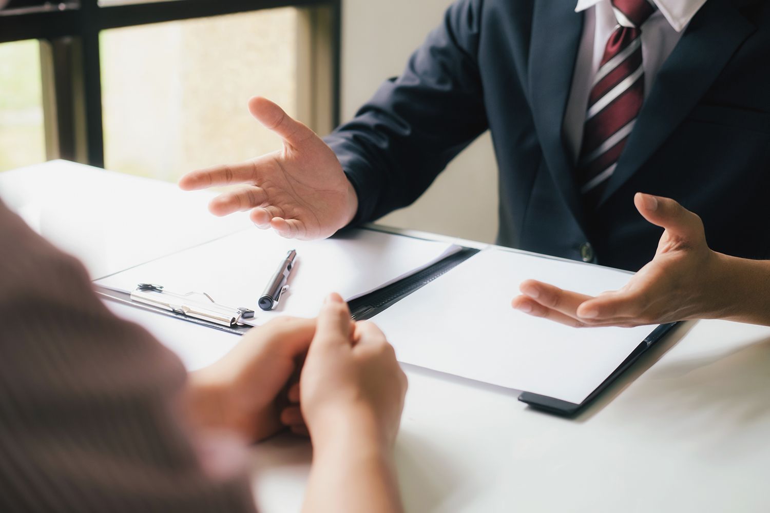 Two people at a desk; one in a suit gestures, the other listens and has hands clasped. Documents and pen visible.
