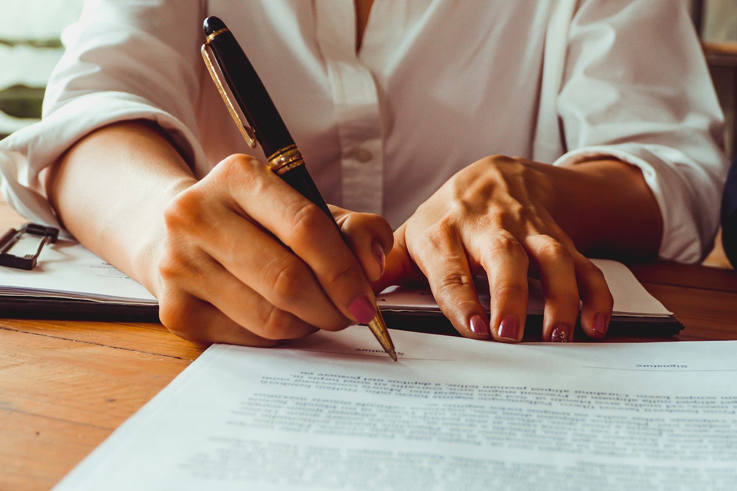 Woman's hands writing with a pen on a document at a wooden table.