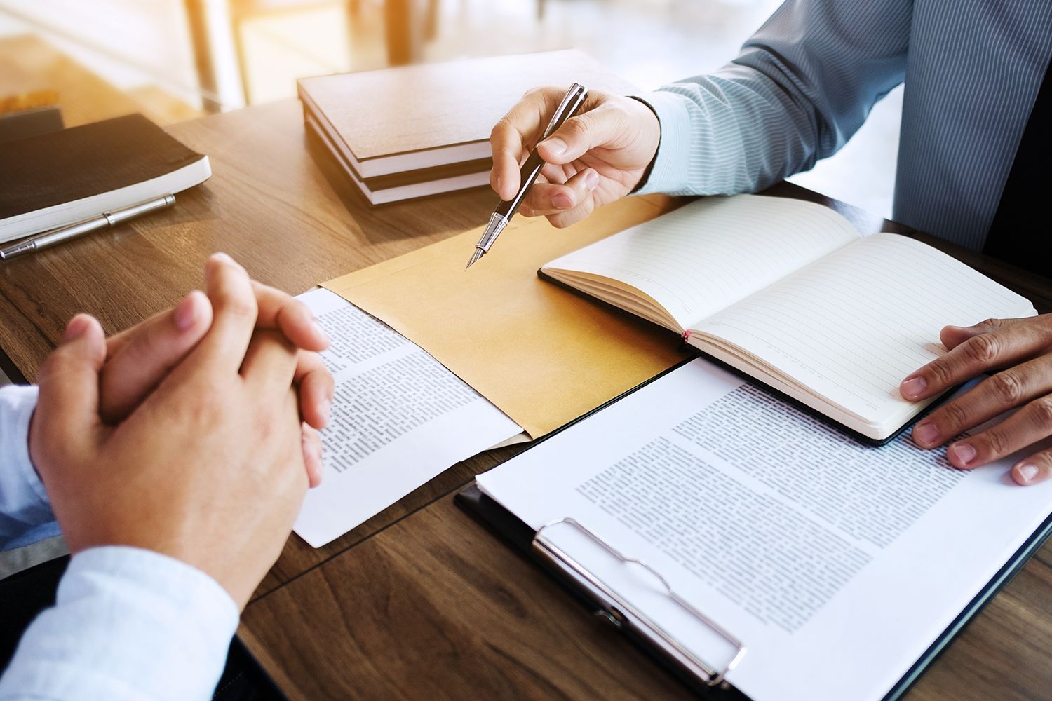 Two people at a desk reviewing documents; one points a pen at a paper while another clasps hands.