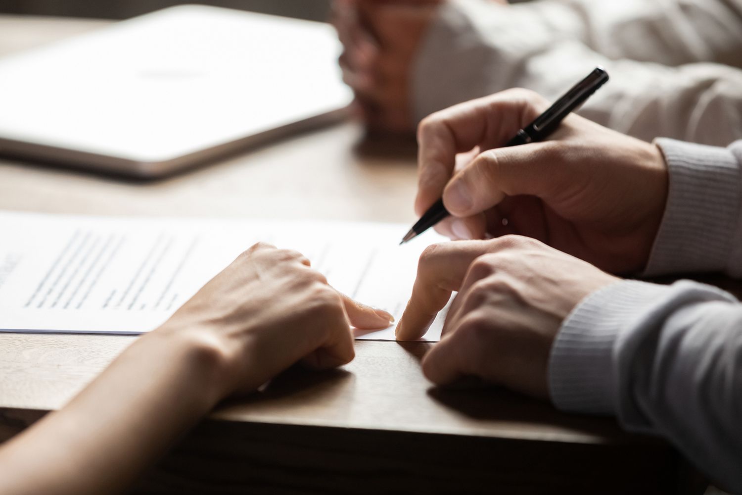 Hands signing a document with a pen, another person pointing at the paper.