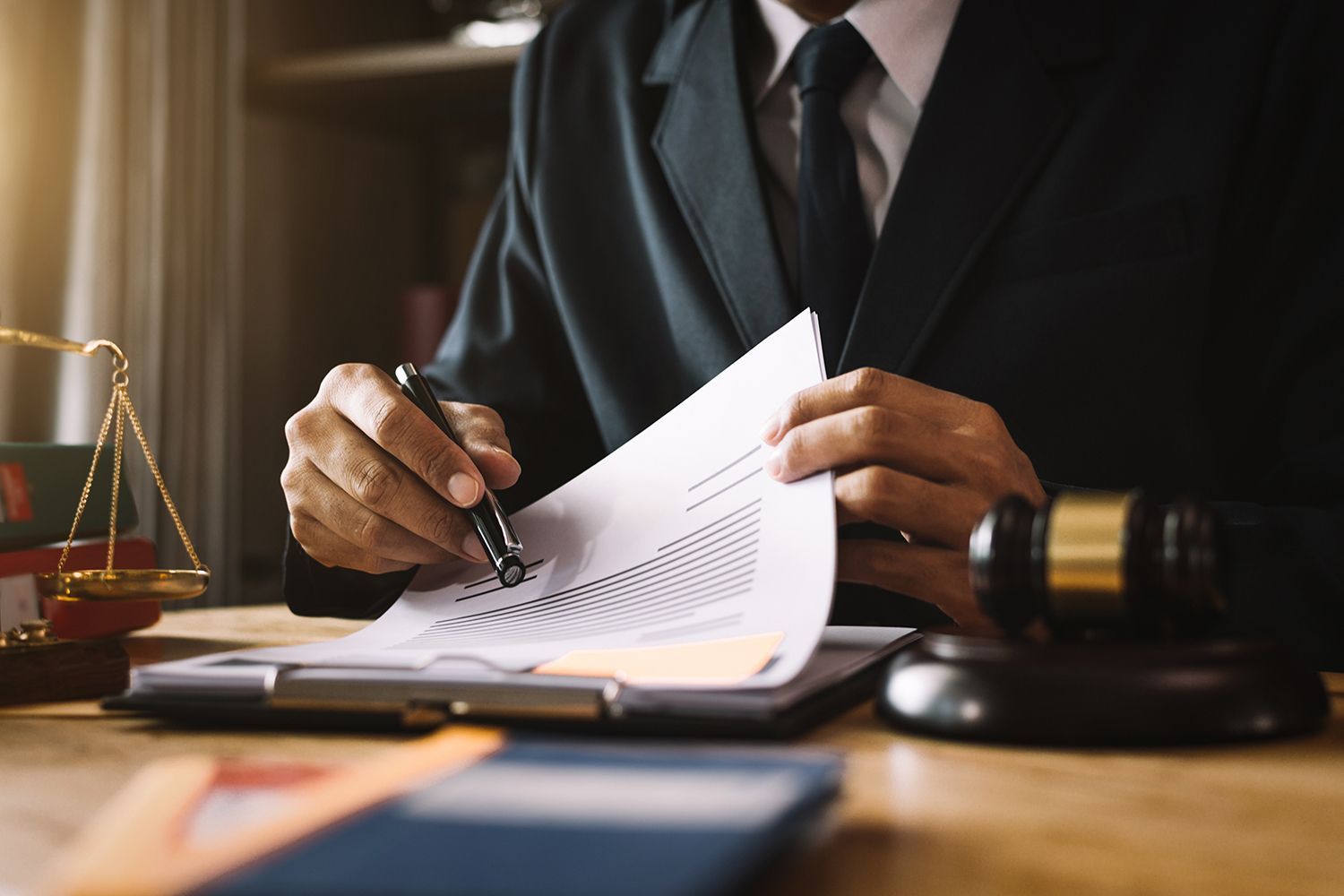 Lawyer signing a document at a desk with a gavel and scales of justice in the background. Lawyer signing a document at a desk with a gavel and scales of justice in the background.
