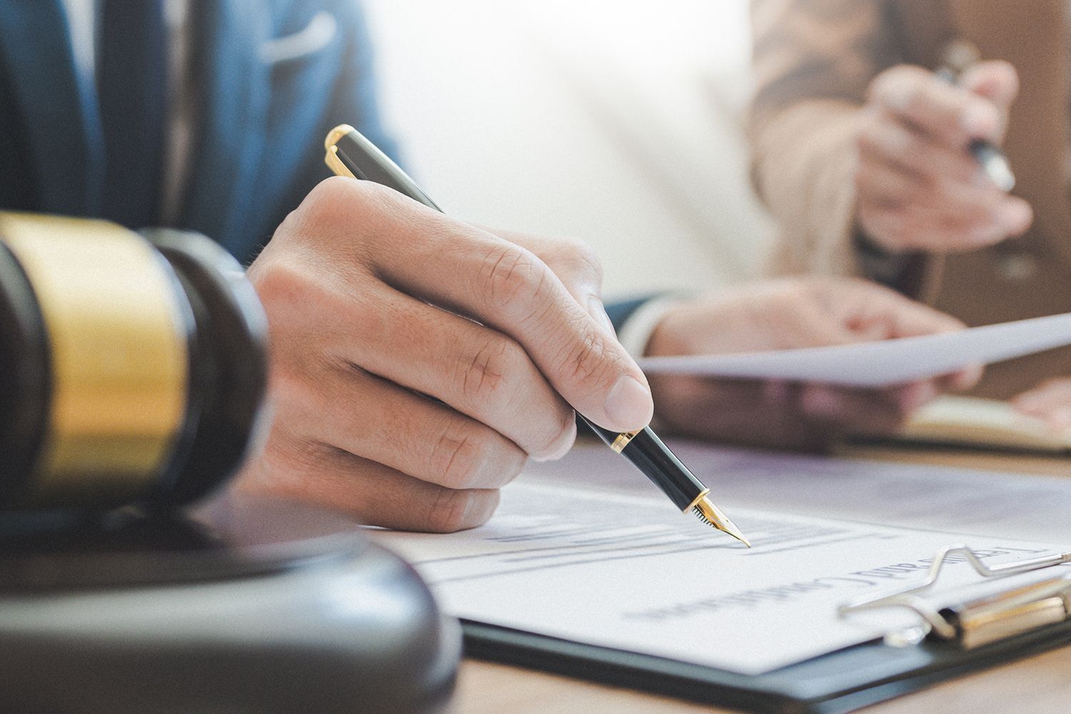 Person signing document with pen, gavel in foreground, other person holding papers.