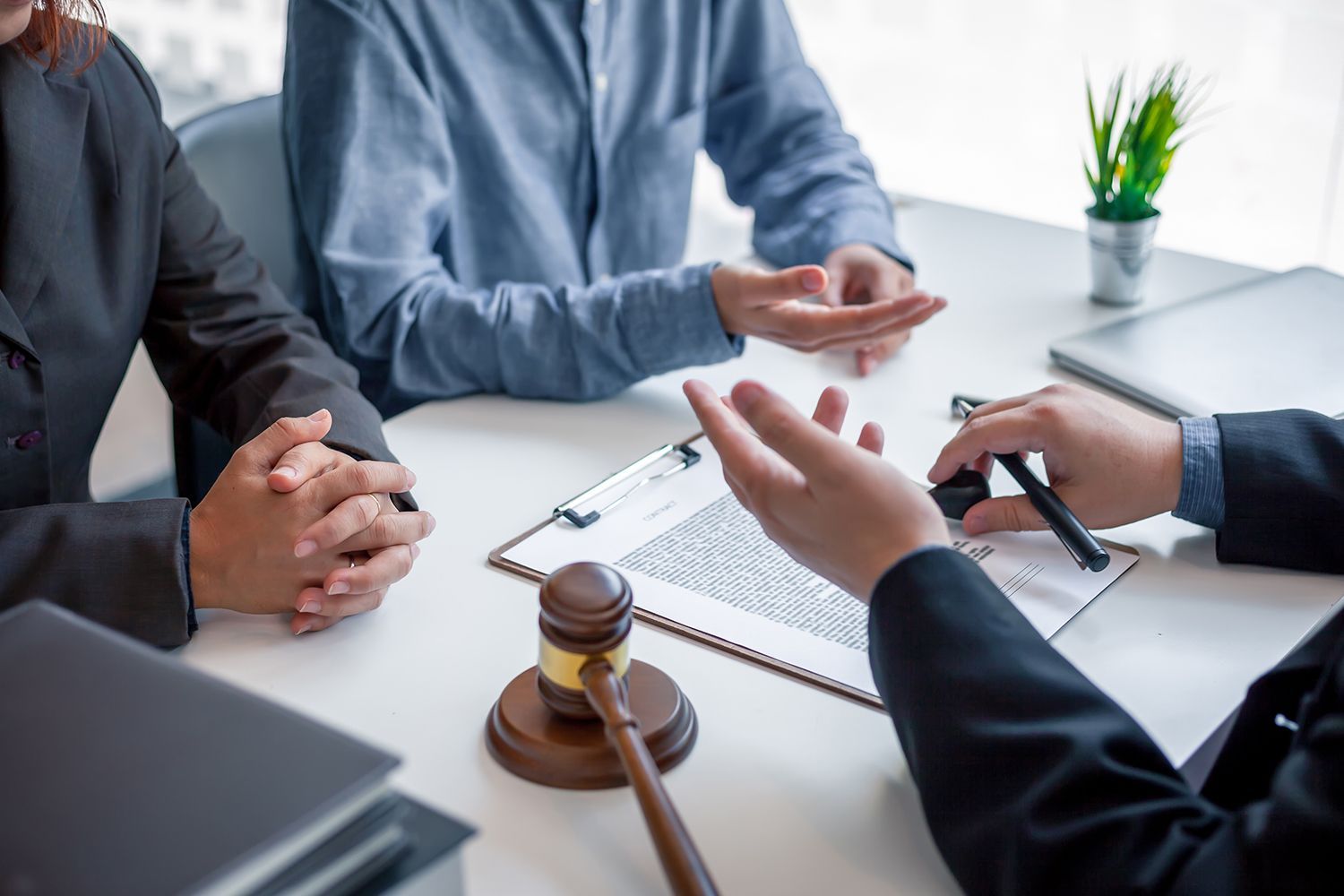 Two people at a desk with a lawyer, discussing documents. Gavel, pen, and a small potted plant on the table.