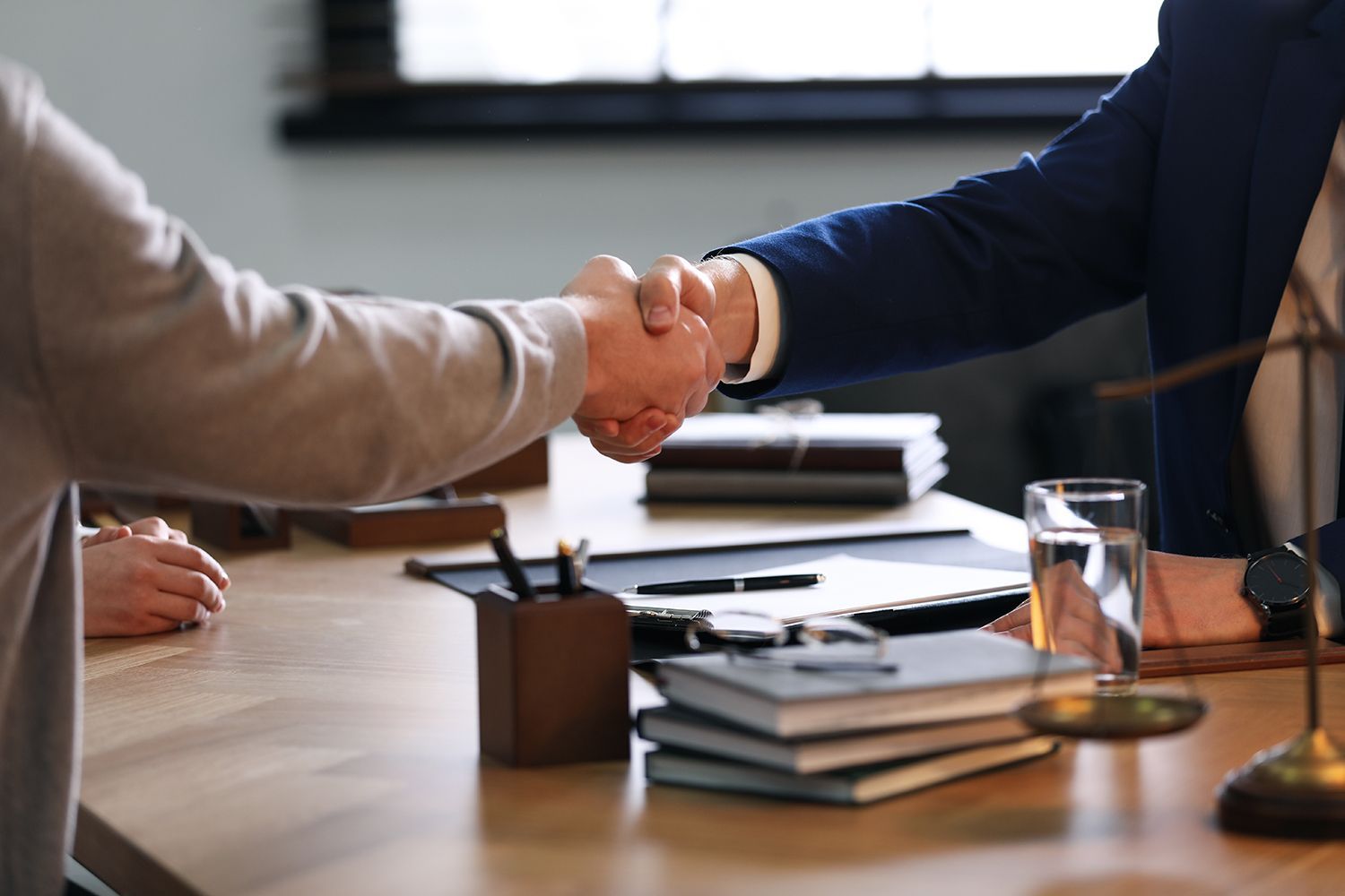 Two people shaking hands over a desk with legal documents and a scale, indoors.
