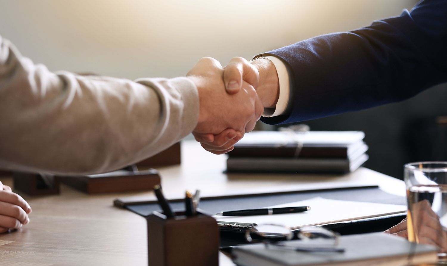 Two men shaking hands over a desk with paperwork, symbolizing an agreement.